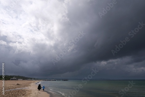 Dramatic scenery with stormy clouds over beach of Baltic Sea and wooden pier in Brzezno, Jelitkowo. Pomerania, Poland. Little silhouettes of woman with child.