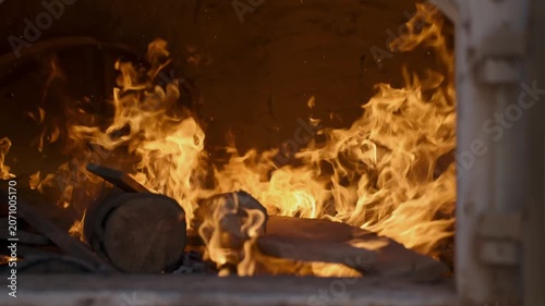 Bright orange and yellow flames dance and roar inside a traditional stone kiln or industrial furnace, consuming wooden logs with intense heat and glowing light in a dark interior space