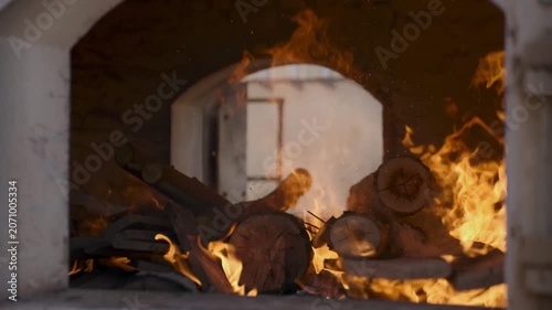 Intense bright orange flames burning dry wooden logs inside a traditional stone brick oven used for baking bread or pizza in a rustic kitchen setting with warm lighting and glowing embers