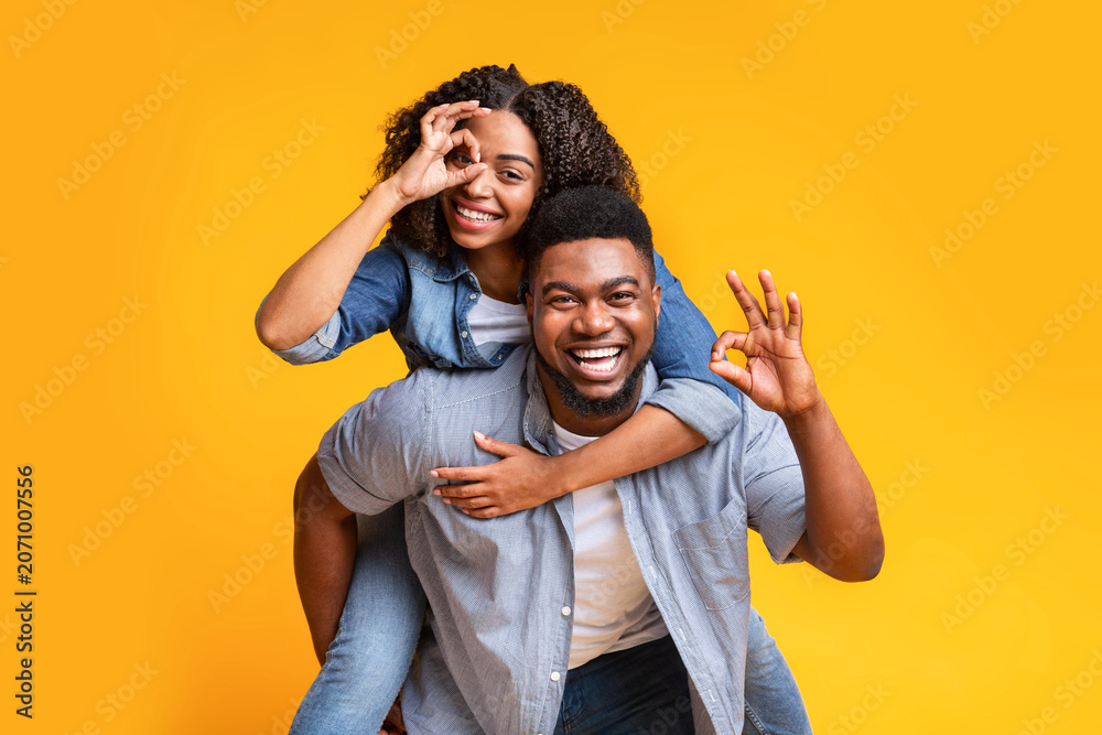 Fototapeta premium Smiling Black Young Couple Enjoying Playful Moment. Man Giving Woman Piggyback Ride While Showing Hand Gestures, Having Fun Together In Studio At Modern Yellow Background. Concept Of Happiness.