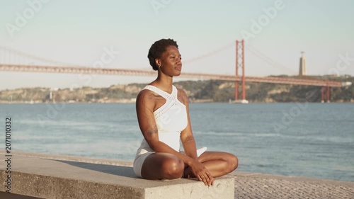 Black woman sitting cross legged near waterfront during sunset while camera slowly zooms in on face. Calm evening atmosphere with bridge and river in background
