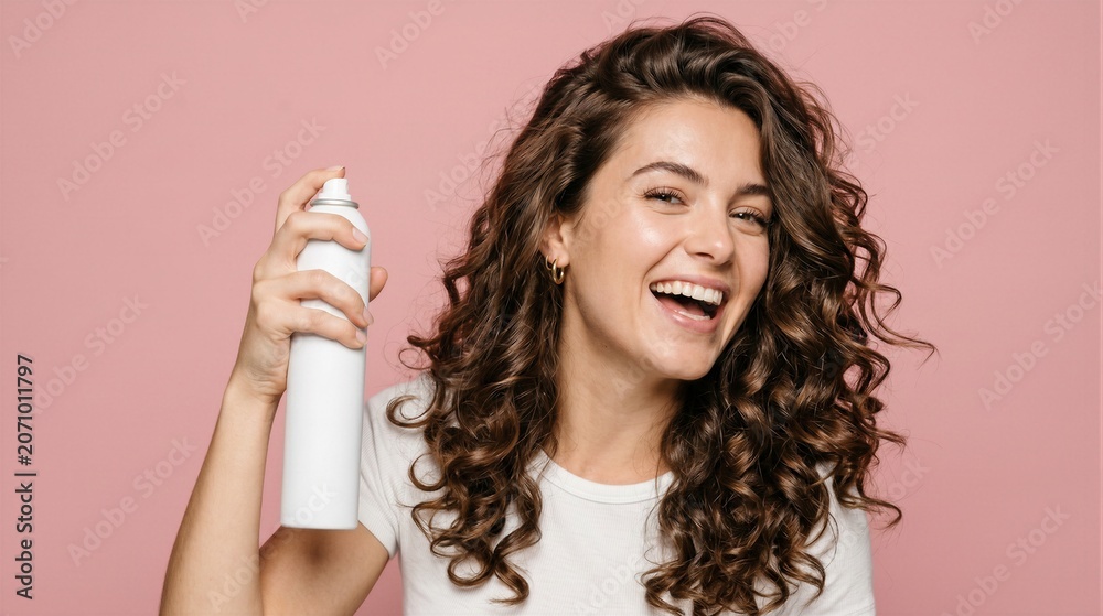 Fototapeta premium Happy woman with curly hair holding a hairspray bottle, isolated on a pink background