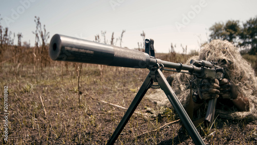 Tableau sur toile Sniper team member in camouflage gear aiming a suppressed rifle from a prone pos