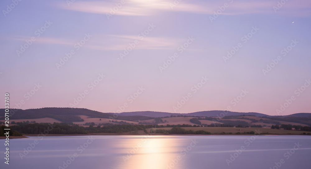 Fototapeta premium A tranquil lake reflects the soft hues of a twilight sky, with rolling hills and distant lights creating a peaceful landscape.