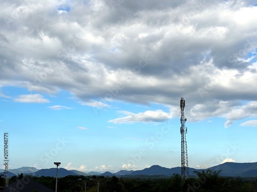 A solar-powered cellular communication and lighting tower stands in a remote landscape surrounded by lush greenery, rolling hills, and bright cloud formations.