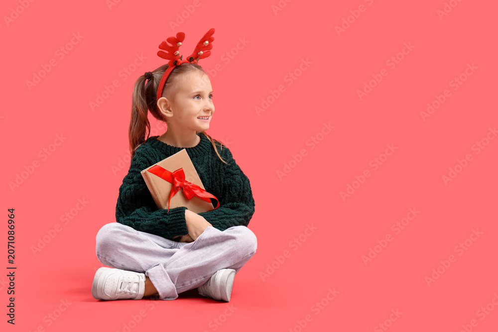 Naklejka premium Cute little girl in Christmas reindeer horns with gift book sitting on red background