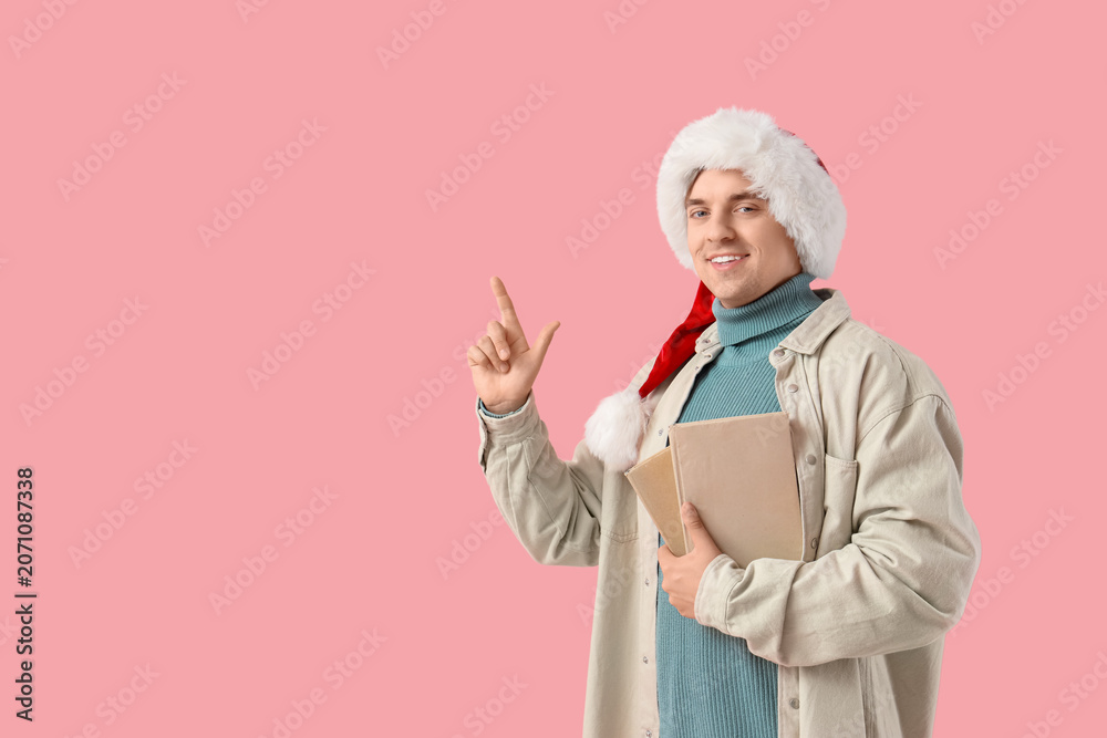 Naklejka premium Young man in Santa hat and with books pointing at something on pink background