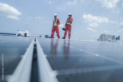 Tablou pe pânză On-site view as pair of technicians in red protective suits stride across rows of photovoltaic panels on a rooftop solar farm, capturing green technology, solar power generation