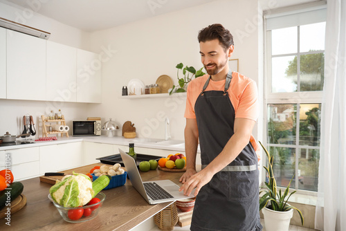 Handsome bearded man with laptop cooking in kitchen
