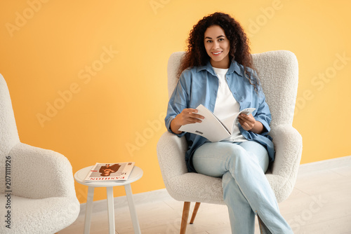 Young African-American woman reading magazine in white armchair near orange wall