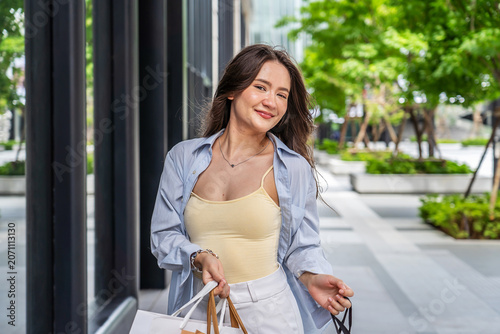 Happy young Asian woman walking on a modern city street holding shopping bags. She smiles and looks at store windows enjoying a weekend shopping trip. Fashionable lifestyle and retail therapy.