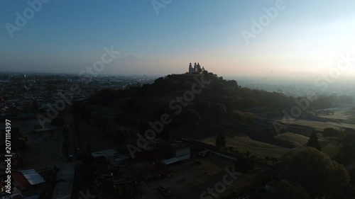Aerial drone moves cinematically close to the Church of Our Lady of Remedies atop the Cholula pyramid at dawn, with the town and Puebla city spreading across the background