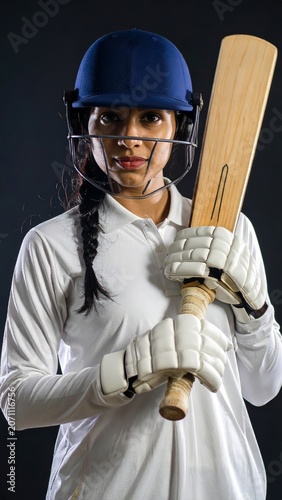 Portrait of a Female Cricket Player in Full Gear Holding a Bat Against a Dark Background