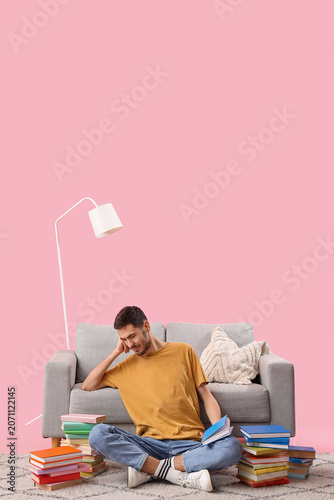 Young man with books sitting near sofa on pink background
