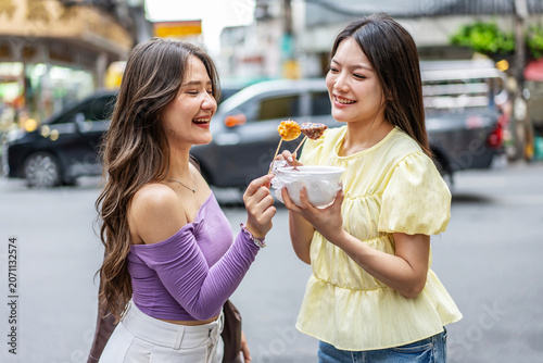 Two happy young Asian women friends enjoying delicious local street food together on a vibrant city street during a vacation trip in Thailand. Joyful lifestyle, travel, and food tourism concepts.