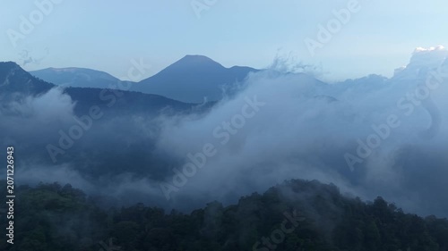 Aerial view of dense forest blanketed in thick mist, with towering evergreen trees partially obscured by fog. Mist creates mysterious atmosphere, while distant mountains barely emerge through haze.
