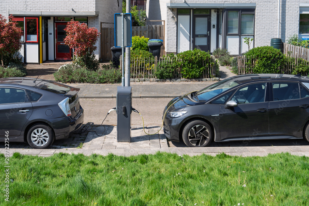 Obraz premium Two modern electric vehicles parked on a quiet residential street in the Netherlands, connected to a public charging station pillar. Sustainable urban transport infrastructure.