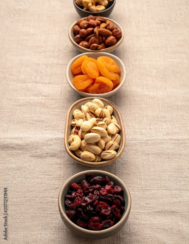 Row of small bowls filled with nuts, dried apricots, and cranberries on a rustic linen texture background for healthy snack concepts