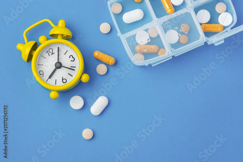 Pills and supplement with yellow alarm clock and medication pill box on blue background, top view