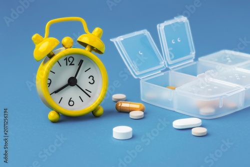 Yellow alarm clock with scattered pills and pill box on blue background, time to take medicine concept