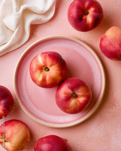 Soft pink plate holds three ripe peaches. Peaches gleam with natural blush and warm tones. Crisp white cloth drapes beside the plate. Scattered peaches enhance the inviting arrangement