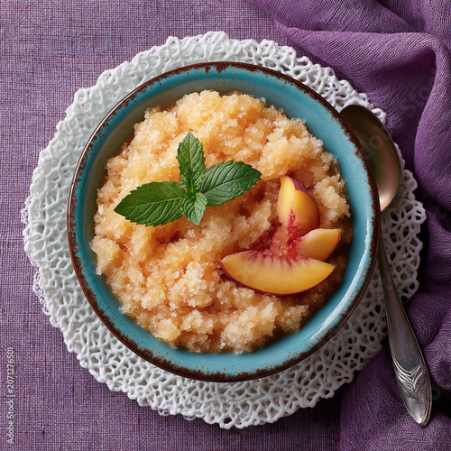Peach granita glisters in a blue bowl. Fresh mint and peach slices garnish the dessert. Delicate lace doily cradles the bowl. Purple fabric adds rich, soothing contrast