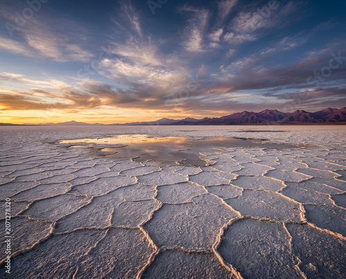 Sunset over a vast salt flat, showcasing intricate ground patterns and a reflective pool mirroring the vibrant sky and distant mountains