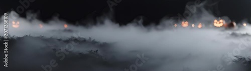 Jack o lantern faces glow in the distance partially obscured by heavy fog and mist during a dark night.