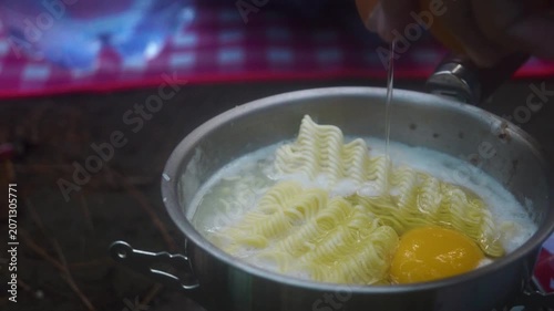 A hand stirs instant noodles and egg into boiling water in a metal bowl during an outdoor camping trip, demonstrating a simple meal preparation method