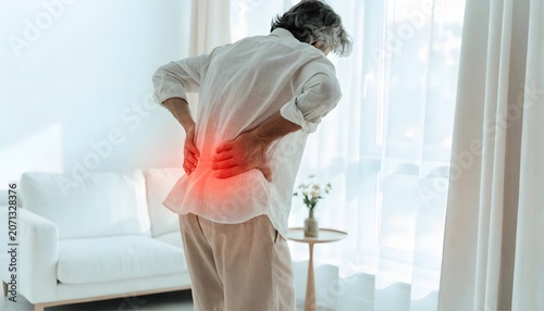 Senior man suffering from acute lower back pain at home. Rheumatism relief and sciatica treatment needs. Elderly person standing in a bright living room holding his aching waist area
