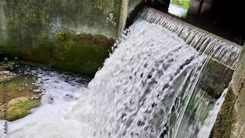 Fast flowing water cascades over a concrete sluice gate, creating foamy currents and dynamic textures. Rustic industrial structure meets nature in a peaceful countryside setting