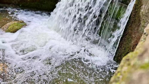 Clear water cascades over a concrete spillway into a foaming stream below. Fast flowing currents, splashing whitewater, mossy rocks, and natural textures create a dynamic outdoor scene