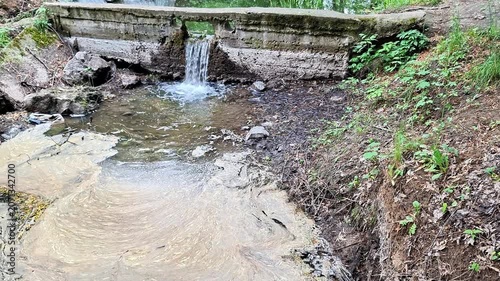 Small waterfall pours from a concrete spillway into a shallow stream surrounded by natural earth banks and greenery. Flowing water, ripples, and peaceful outdoor scenery create a calm atmosphere