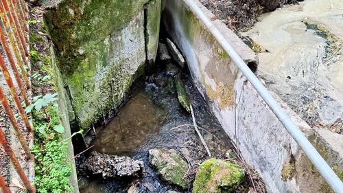 Water flows through a narrow concrete channel beside moss covered stone walls and natural rock formations. Gentle currents, textured surfaces, and a peaceful outdoor setting create a calm countryside 
