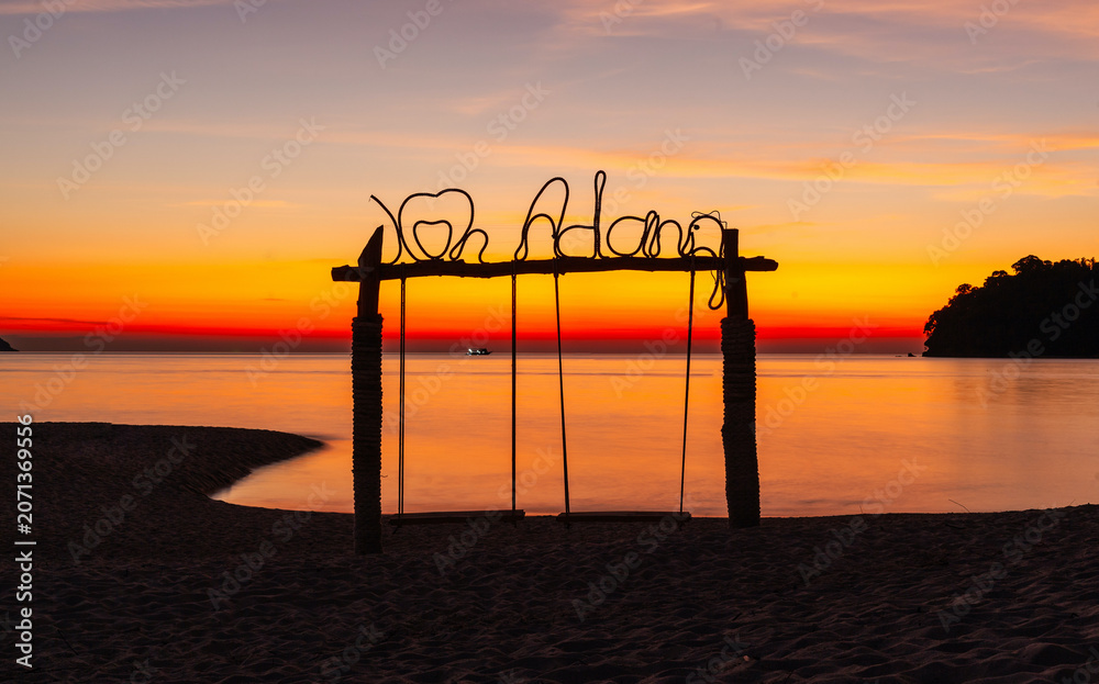 Fototapeta premium Sunset view with Koh Adang sign on beach. Calm ocean waters and vibrant sky. Silhouette of swings and tropical island landscape. Thailand coastal scenery at dusk.