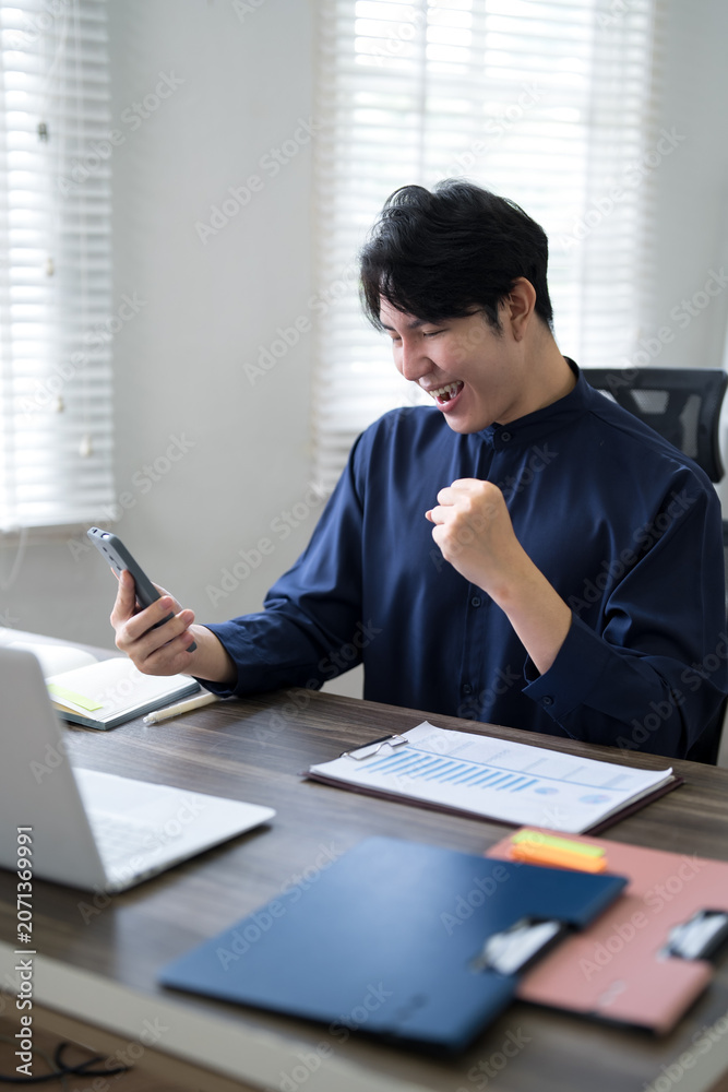 Fototapeta premium Young Asian businessman celebrating success with fist pump while reading smartphone message at modern office desk