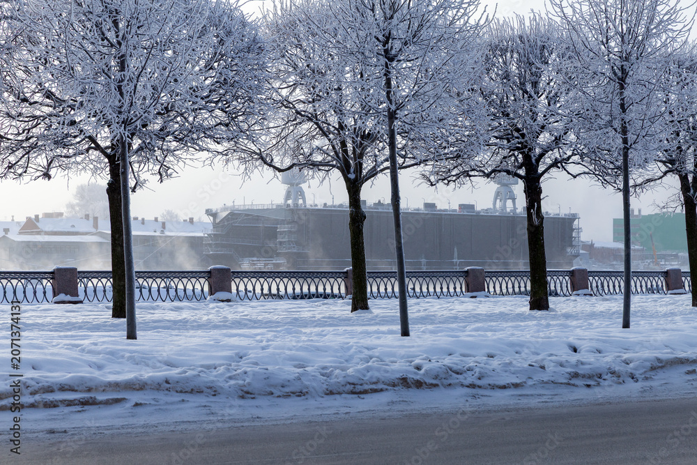 Fototapeta premium A tranquil winter scene along a snow-covered riverside. Bare trees line a frosty path. Saint-Petersburg, Russia