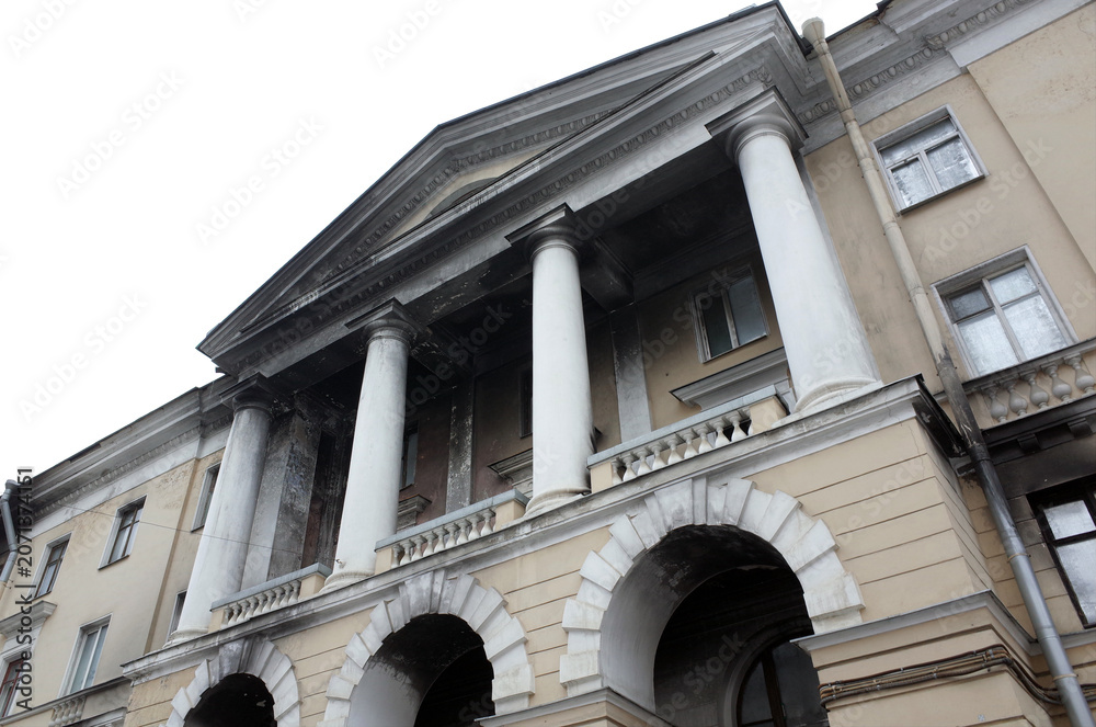 Naklejka premium Neoclassical building facade after a fire photographed from a low angle, showing tall columns, arched entrances, balustrade balcony and weathered plaster that conveys history, grandeur and urban decay