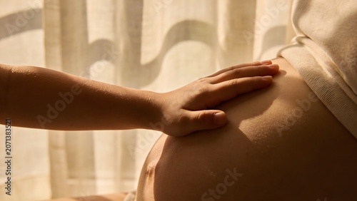 Close-up of a child's hand touching a pregnant mother's belly in warm sunlight