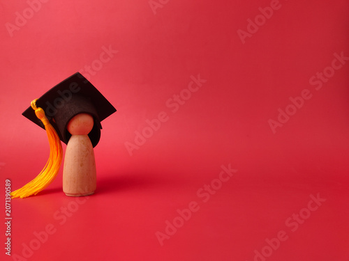 A single wooden peg figure stands on a red background wearing a miniature black graduation cap with a yellow tassel