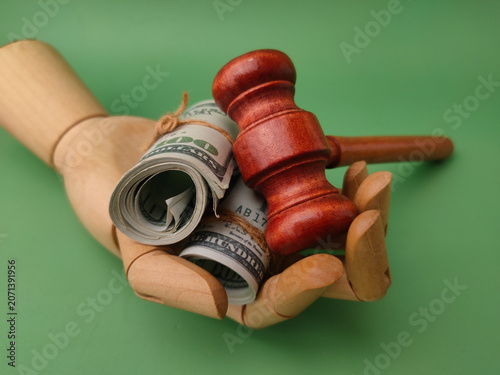 A wooden mannequin hand holds two tied rolls of dollar bills and a wooden judge gavel against a green background
