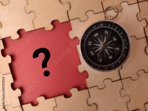 A circular magnetic compass rests on a wooden jigsaw puzzle next to a missing piece showing a question mark