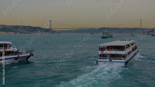 Two white passenger ferries cruising on the blue waters of the Bosphorus in Istanbul with the iconic suspension bridge and city skyline under a soft sunset sky
