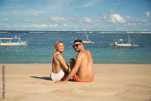 Couple relaxes on sandy beach while facing the ocean and boats