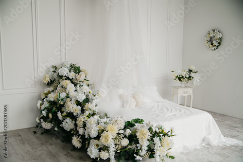 Interior of a photo studio painted in white colors, with a large sofa covered with white blankets, surrounded by white roses and a transparent tulle hanging above it