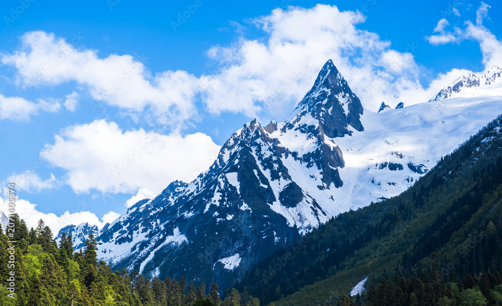 Obraz premium Majestic snow-capped mountains tower against a clear blue sky with clouds in Dombay, Russia, highlighting the region's breathtaking landscape