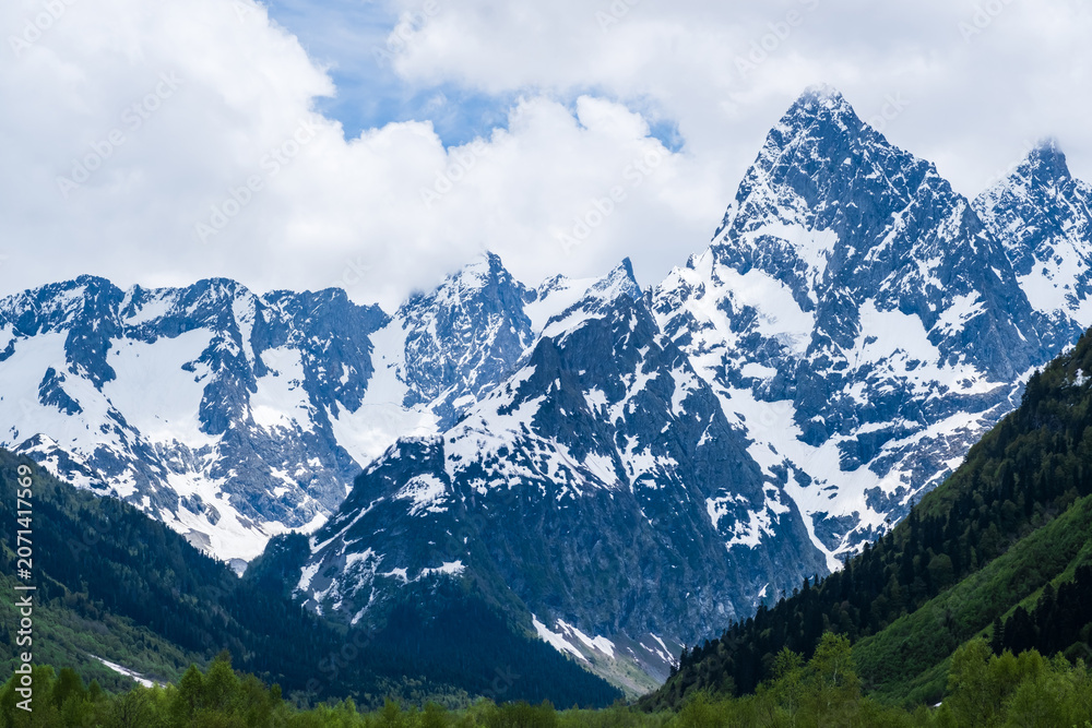 Obraz premium Snow-covered mountain peaks tower against a cloudy sky, with green valleys and rocky terrain creating a stunning natural landscape