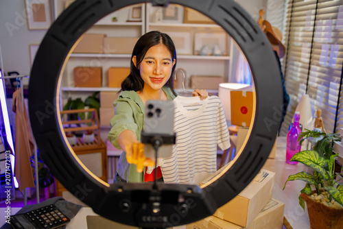 Young Asian woman fashion entrepreneur live streaming selling clothes online with smartphone ring light and laptop in home studio, representing small business owner and professional women.