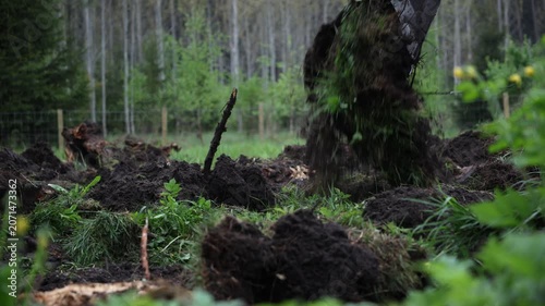 Excavator bucket digs into dark turf, flinging soil and roots across a fenced rural woodlot near birch like trees, with stumps and sod piles on an overcast day.