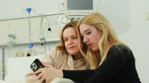 Mother and daughter lean close for smartphone selfie in hospital bed. Blond daughter extends phone forward framing lovely faces for photo